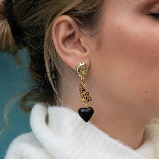 Close-up of a person wearing gold earrings with a black heart charm against a blue background