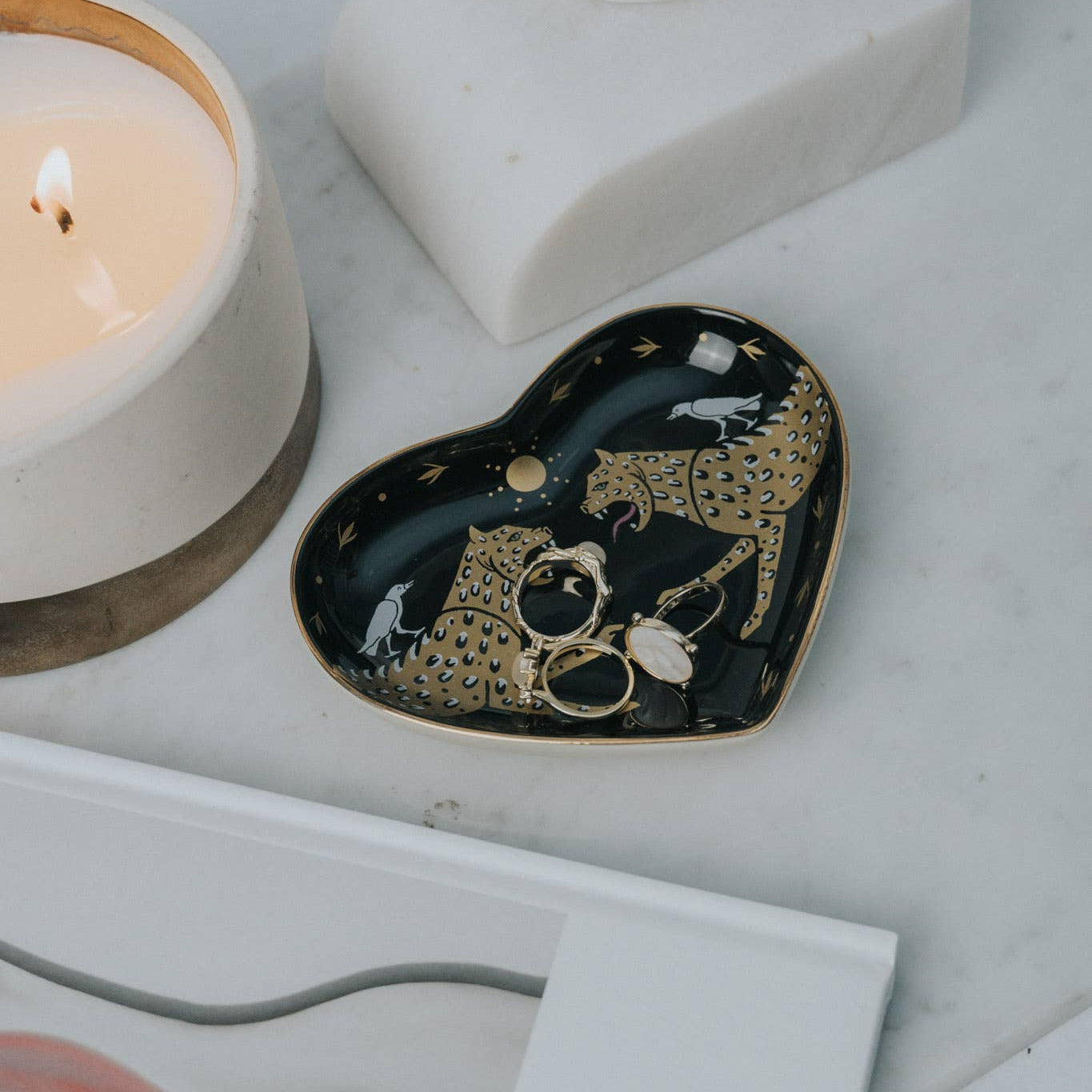 Heart-shaped decorative tray with gold accents on a marble surface next to a candle and sculpture.