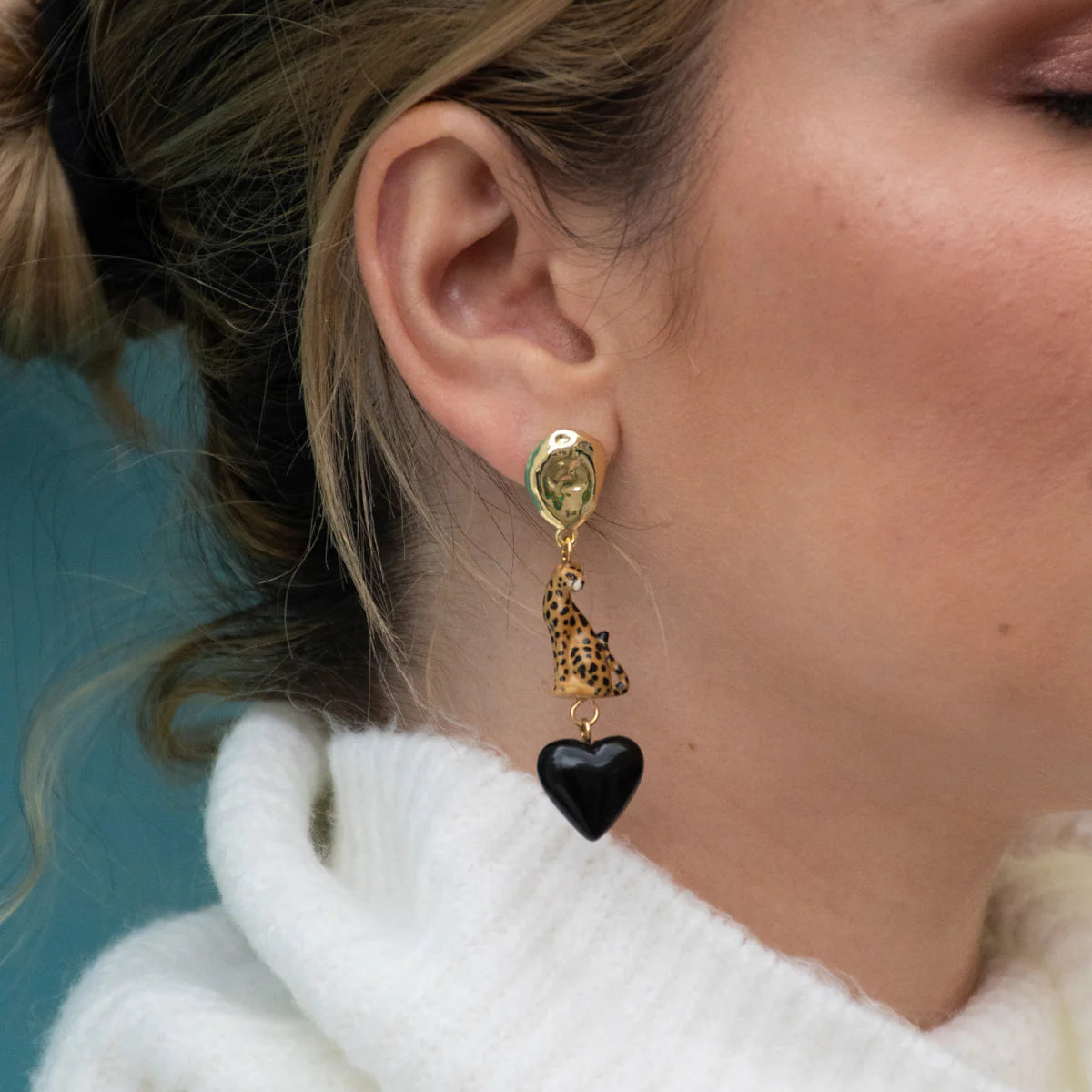 Close-up of a person wearing gold earrings with a black heart charm against a blue background