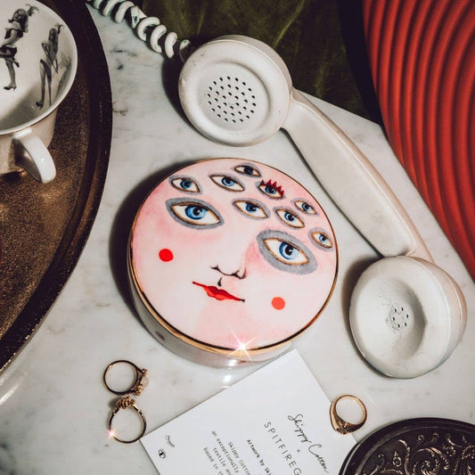 Trinket box with artistic face design on a marble surface with a vintage phone and other items.