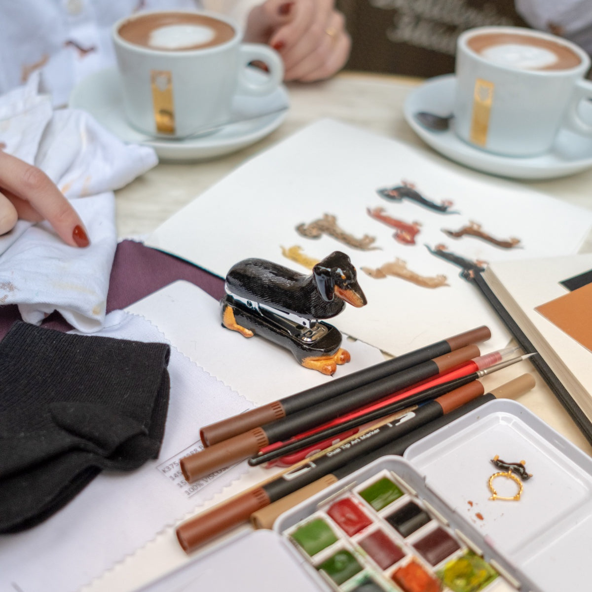 Desk with stationery items, including pens, a stapler, and watercolor set, with people sitting around a table with coffee.